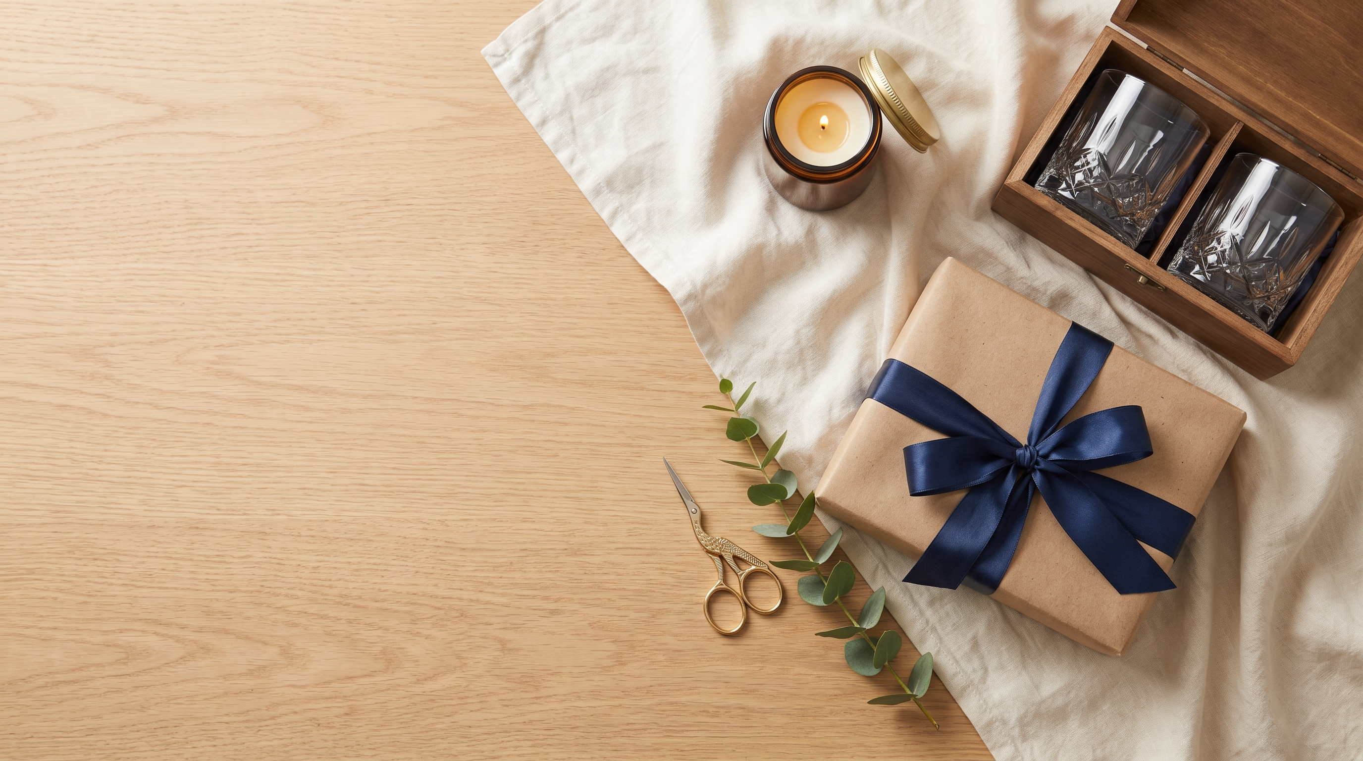 Geschenke einpacken von oben: Kraftpapier-Paket mit navyblauem Band, Messingschere und Whiskyglaeser in Geschenkbox auf Eichentisch